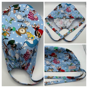 May include: A blue and white surgical scrub hat with a white polka dot pattern. The hat features a print of characters from Alice in Wonderland, including the Mad Hatter, the Queen of Hearts, and the Cheshire Cat. The hat has a tie at the back and the text "We're all mad here" is printed on the hat.
