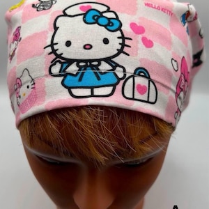 May include: Pink and white patterned scrub hat with a Hello Kitty design. The hat features a pink and white checkered pattern with hearts and Hello Kitty characters. The main character is a nurse Hello Kitty with a blue dress and a white hat. The text "Hello Kitty" is printed on the hat.