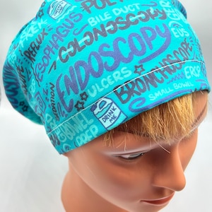 May include: Turquoise blue surgical scrub hat with a white seam. The hat is covered in a colorful print of medical terms, including "Endoscopy", "Bronchoscopy", "Colonoscopy", "Esophagus", "Polyps", "Ulcers", "Small Bowel", and "Drink Me".