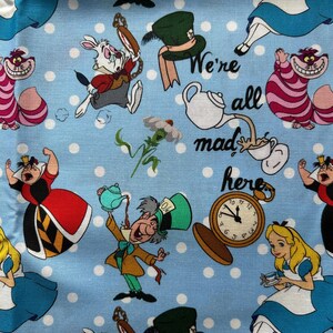 May include: A blue fabric with white polka dots features a whimsical Alice in Wonderland theme. The fabric includes characters like the Mad Hatter, the Queen of Hearts, the Cheshire Cat, and Alice herself. The text "We're all mad here" is also included.