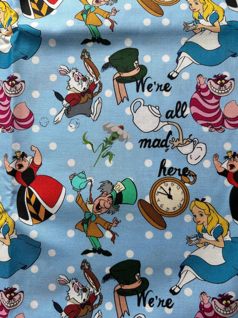 May include: A blue fabric with white polka dots features a whimsical Alice in Wonderland theme. The fabric includes characters like the Mad Hatter, the Queen of Hearts, the Cheshire Cat, and Alice herself. The text "We're all mad here" is also included.
