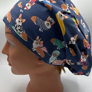 May include: A blue scrub hat with a pattern of corgis wearing different outfits. The hat has a white seam around the edge and a small white tag with the text "Scrub Caps by Shawna" on the back.