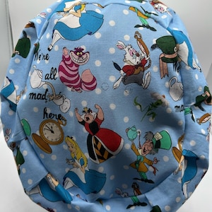 May include: A blue fabric scrub hat with a white polka dot pattern and a repeating print of characters from Alice in Wonderland. The text "We're all mad here" is printed on the hat.
