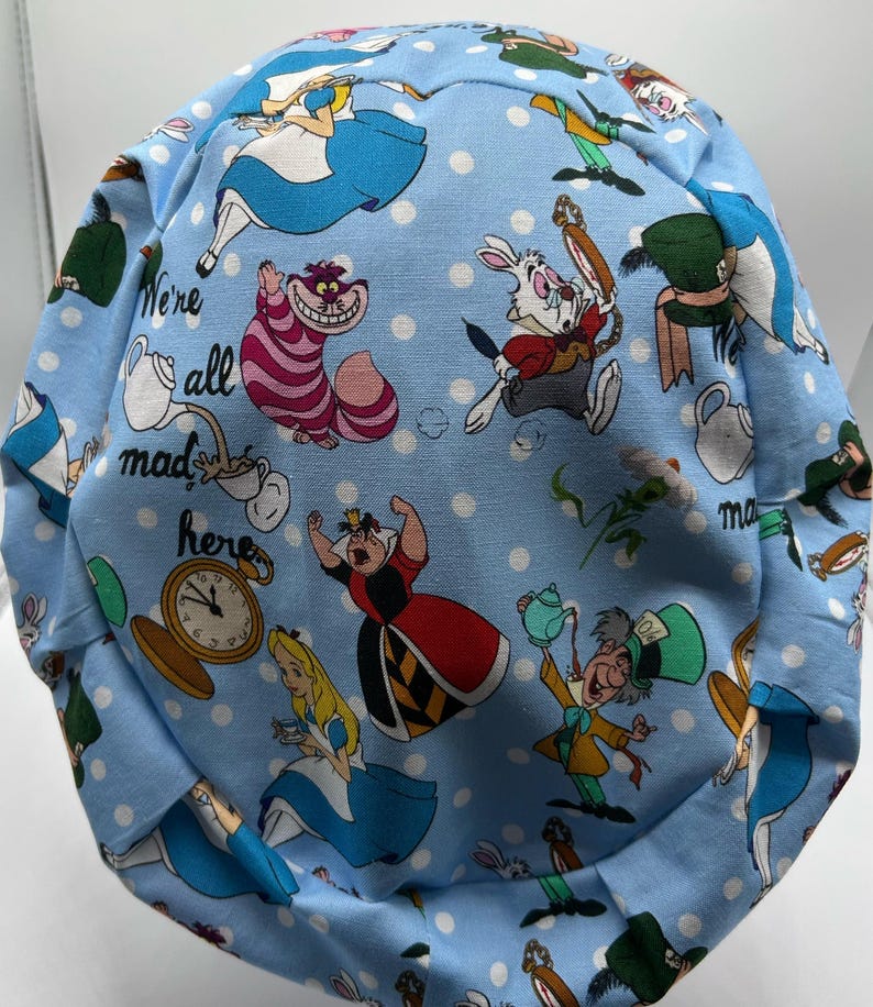 May include: A blue fabric scrub hat with a white polka dot pattern and a repeating print of characters from Alice in Wonderland. The text "We're all mad here" is printed on the hat.