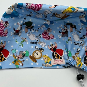 May include: Blue scrub hat with a white polka dot pattern and a colorful Alice in Wonderland print. The print features characters like the Queen of Hearts, the Mad Hatter, and the Cheshire Cat. The hat has a drawstring closure and the text "We're all mad here" is printed on the hat.