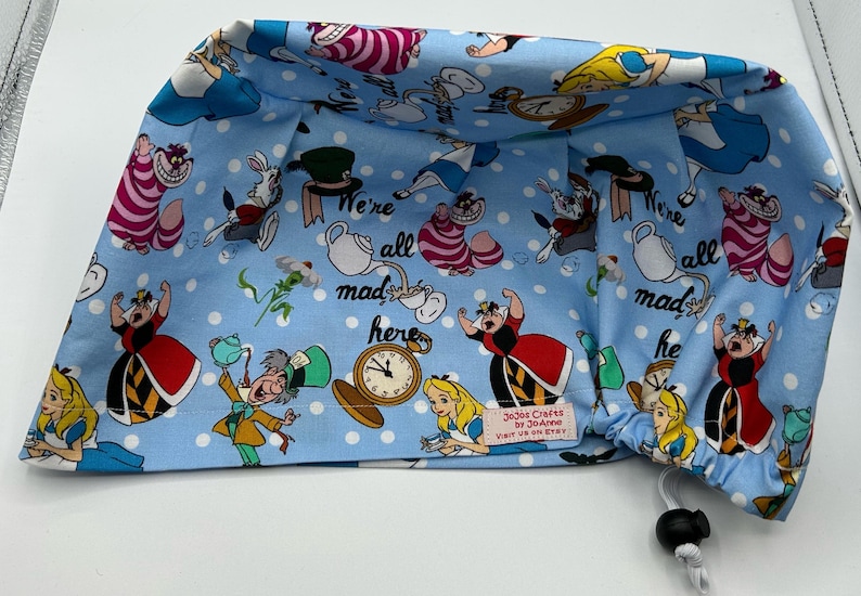 May include: Blue scrub hat with a white polka dot pattern and a colorful Alice in Wonderland print. The print features characters like the Queen of Hearts, the Mad Hatter, and the Cheshire Cat. The hat has a drawstring closure and the text "We're all mad here" is printed on the hat.