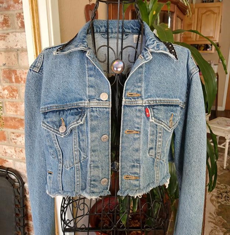 Op de afbeelding: Een lichtblauwe spijkerjas met een korte, gerafelde zoom. De jas heeft twee borstzakken, knoopsluitingen en een klein rood label. De kraag heeft zwart-witte stippenaccenten. Een klassiek, casual bovenkledingstuk.