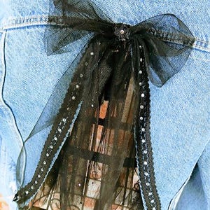 Op de afbeelding: Een zwarte tule strik met strass accenten is bevestigd aan de achterkant van een lichtblauwe spijkerjas. De strik is gecentreerd en voegt een decoratief element toe aan de jas. De tule heeft een transparante kwaliteit.