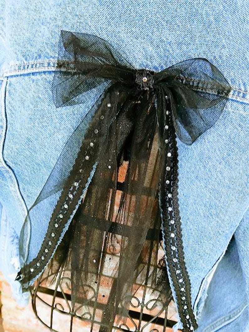 Op de afbeelding: Een zwarte tule strik met strass accenten is bevestigd aan de achterkant van een lichtblauwe spijkerjas. De strik is gecentreerd en voegt een decoratief element toe aan de jas. De tule heeft een transparante kwaliteit.
