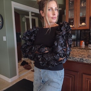 May include: Black top with decorative floral sleeves. The sleeves are made of a dark, textured fabric, creating a dramatic effect. The top is paired with a denim skirt. The background includes a kitchen and a dartboard.