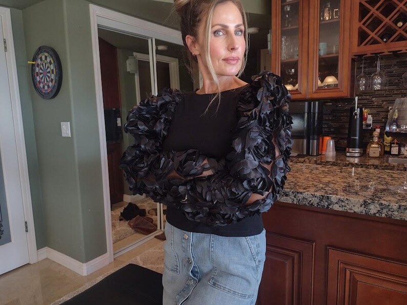 May include: Black top with decorative floral sleeves. The sleeves are made of a dark, textured fabric, creating a dramatic effect. The top is paired with a denim skirt. The background includes a kitchen and a dartboard.