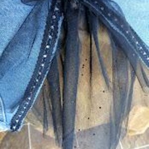Op de afbeelding: Een zwarte tule strik met een decoratieve zilveren rand en een sprankelende, doorschijnende overlay. De strik is bevestigd aan een blauwe spijkerbroek. Het ontwerp van de strik omvat lange, vloeiende linten.