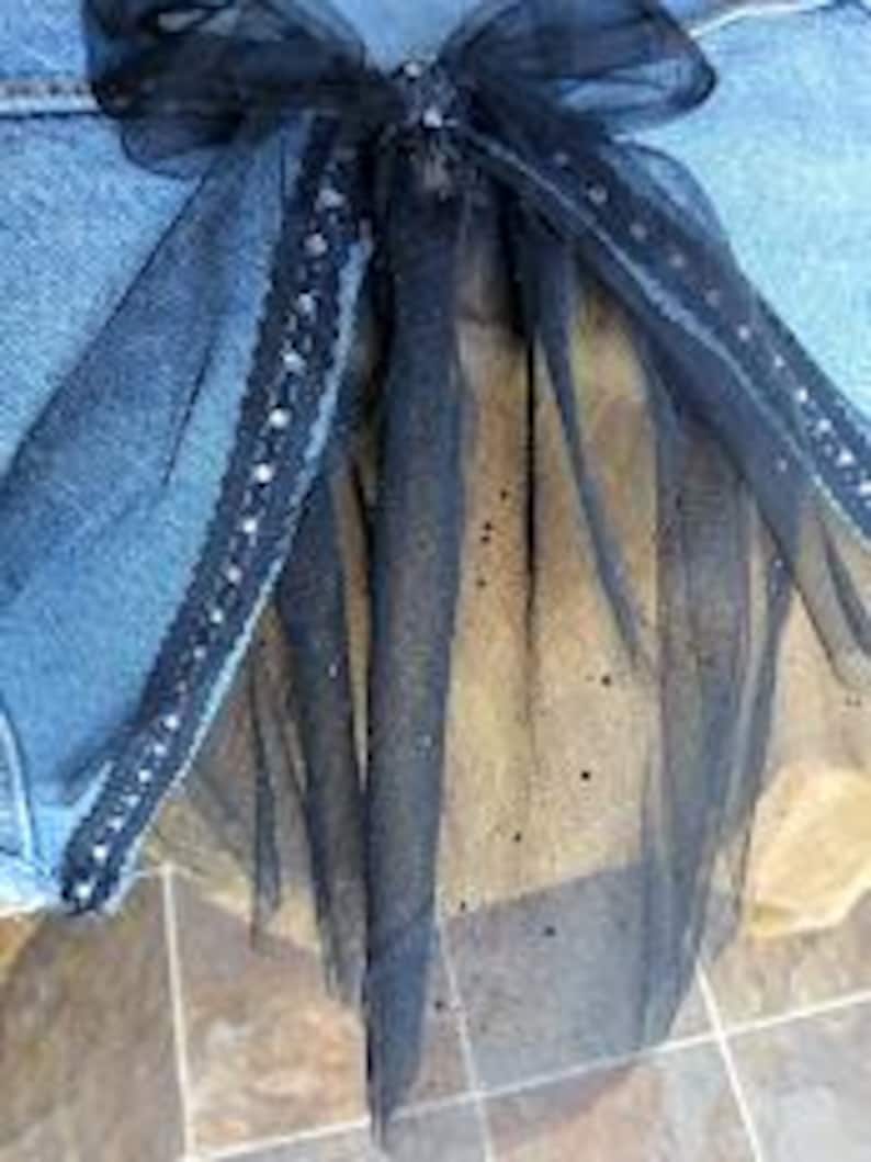 Op de afbeelding: Een zwarte tule strik met een decoratieve zilveren rand en een sprankelende, doorschijnende overlay. De strik is bevestigd aan een blauwe spijkerbroek. Het ontwerp van de strik omvat lange, vloeiende linten.