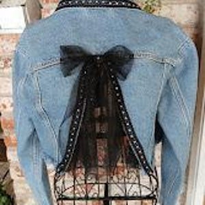 Op de afbeelding: Lichtblauwe spijkerjas met een zwarte tule strik en decoratieve afwerking op de rug. De jas heeft een klassieke kraag, een knoopsluiting aan de voorkant en lange mouwen. De achterkant heeft een zwarte tule strik met een decoratieve afwerking.