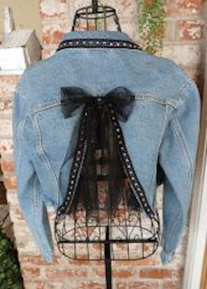 Op de afbeelding: Lichtblauwe spijkerjas met een zwarte tule strik en decoratieve afwerking op de rug. De jas heeft een klassieke kraag, een knoopsluiting aan de voorkant en lange mouwen. De achterkant heeft een zwarte tule strik met een decoratieve afwerking.