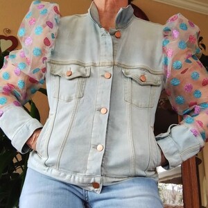 May include: A light blue denim jacket with puffed sleeves made of sheer pink and blue polka dot fabric. The sleeves have a sparkly finish.