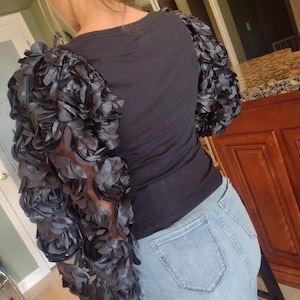May include: Black t-shirt with dramatic, layered, dark gray floral sleeves. The sleeves are the focal point, creating a unique, textured look. The shirt is paired with light blue denim jeans.