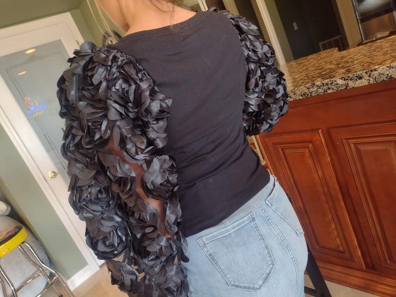 May include: Black t-shirt with dramatic, layered, dark gray floral sleeves. The sleeves are the focal point, creating a unique, textured look. The shirt is paired with light blue denim jeans.