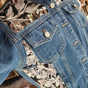 May include: A denim jacket with floral embroidery. The jacket is a medium wash blue with embroidered floral accents in shades of beige and black. It has two chest pockets and button closures.