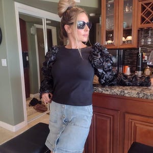 May include: A black long-sleeve top with floral-like embellishments on the sleeves. The person is wearing a denim skirt and sunglasses. A dartboard is visible on the wall. A glass of red wine sits on a countertop.