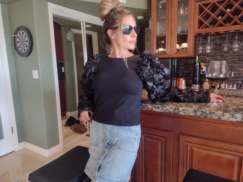 May include: A black long-sleeve top with floral-like embellishments on the sleeves. The person is wearing a denim skirt and sunglasses. A dartboard is visible on the wall. A glass of red wine sits on a countertop.