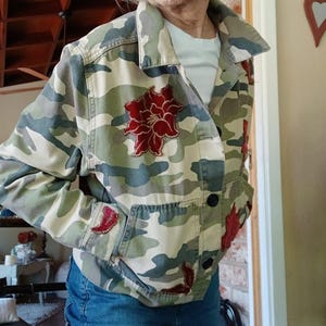 Könnte beinhalten: Eine Tarnjacke mit roter Samt-Blumen Stickerei. Die Jacke hat einen Knopfverschluss und eine Tasche auf der linken Seite.