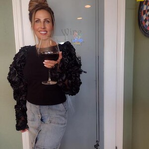 May include: A woman holding a glass of red wine. She is wearing a black top with ruffled sleeves, a denim skirt, and black boots. The background includes a glass door with the word "Wine" and a dartboard.