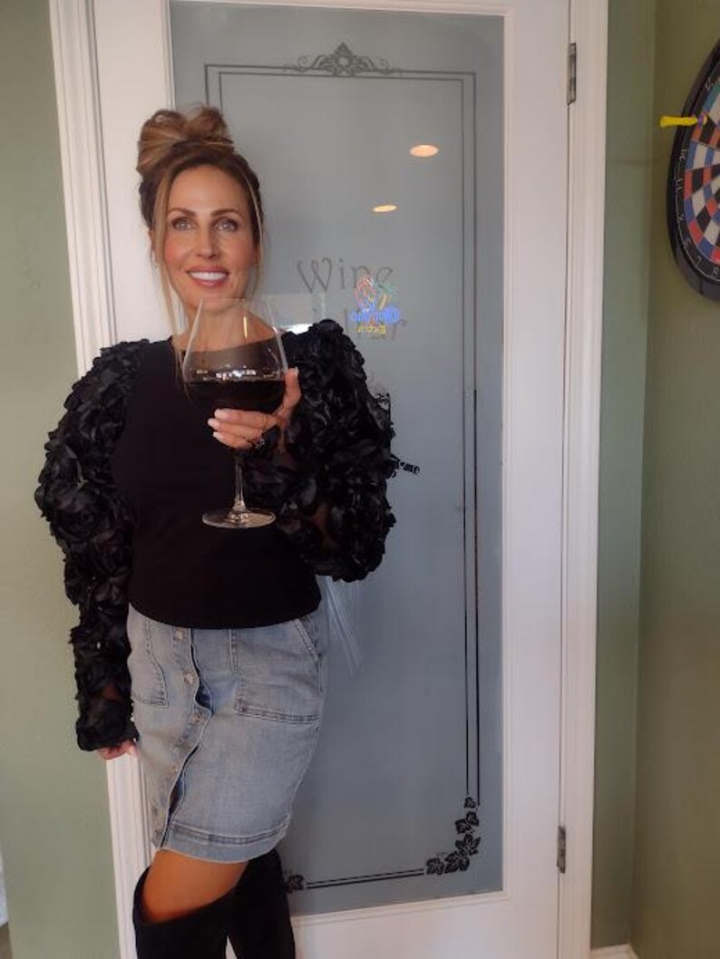 May include: A woman holding a glass of red wine. She is wearing a black top with ruffled sleeves, a denim skirt, and black boots. The background includes a glass door with the word "Wine" and a dartboard.