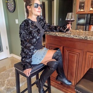 May include: A woman wearing a black floral-textured jacket, denim skirt, and black boots, seated on a black stool. She is holding a glass of red wine. A dartboard hangs on the wall in the background.