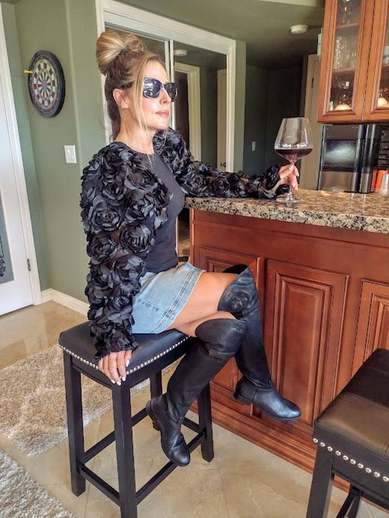 May include: A woman wearing a black floral-textured jacket, denim skirt, and black boots, seated on a black stool. She is holding a glass of red wine. A dartboard hangs on the wall in the background.
