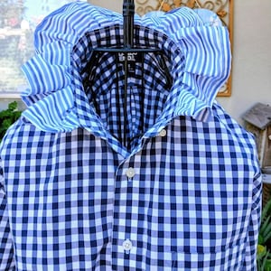 May include: A long-sleeve button-down shirt featuring a navy blue and white gingham check pattern. The shirt includes a large, prominent collar with a blue and white vertical striped ruffle trim and a single chest pocket on the front.