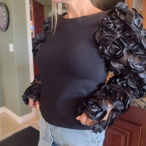 May include: Black long-sleeve top with unique sleeves adorned with black fabric roses. The round-neck shirt is made of a soft material and is paired with blue jeans. The sleeves are the focal point, adding a touch of elegance.
