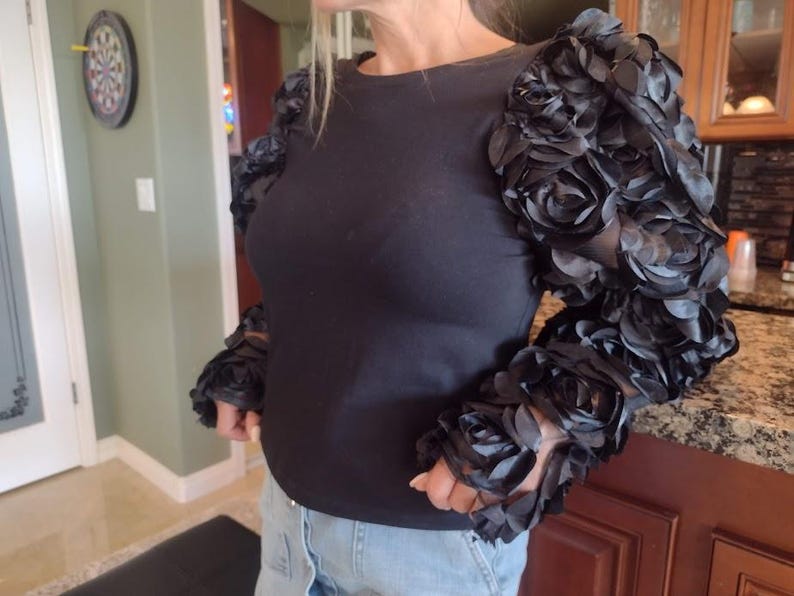 May include: Black long-sleeve top with unique sleeves adorned with black fabric roses. The round-neck shirt is made of a soft material and is paired with blue jeans. The sleeves are the focal point, adding a touch of elegance.