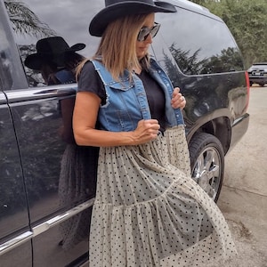 May include: A woman wearing a black cowboy hat, sunglasses, a black t-shirt, a denim vest, and a white tulle skirt with black polka dots. She is standing next to a black SUV.