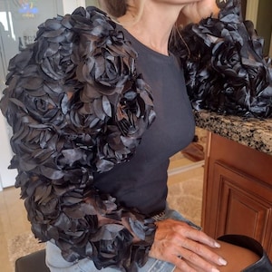 May include: Black, floral-textured bolero jacket with long sleeves. The sleeves are adorned with layered, petal-like fabric, creating a voluminous, rose-like design. The jacket is paired with a dark, form-fitting top.