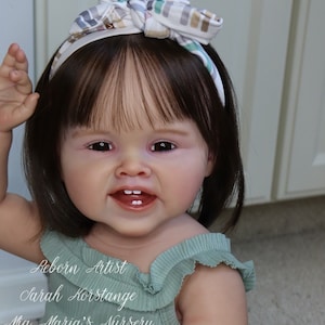 May include: A reborn doll with brown hair and brown eyes wearing a green and white outfit. The doll is sitting on a light brown surface. The text "Reborn Artist Sarah Korstange Ma Maria's Nursery" is visible in the image.