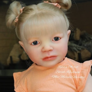 Puede incluir: Primer plano de una muñeca reborn con cabello rubio peinado en coletas con lazos rosas. La muñeca lleva una camisa de color melocotón con el texto "Reborn Artist Sarah Korstange Mia Maria's Nursery".