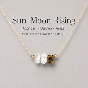 May include: A gold chain necklace featuring three gemstone beads: moonstone, howlite, and tiger eye. Small gold beads flank the gemstones. The text "Sun-Moon-Rising" is above the necklace, with astrological signs and stone names.
