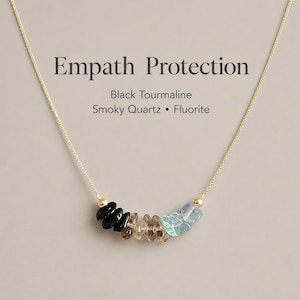 May include: A gold chain necklace featuring a row of small, polished gemstone chips in black, brown, and light blue. The text "Empath Protection" is printed above the stones, followed by "Black Tourmaline, Smoky Quartz, Fluorite".
