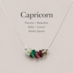 May include: A silver-toned snake chain necklace featuring five small, irregular tumbled gemstones in shades of pale green, deep green, red, dark maroon, and translucent grey. Text above reads: Capricorn, Fluorite, Malachite, Ruby, Garnet, Smoky Quartz.