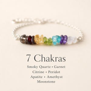 May include: A silver chain bracelet featuring seven colorful gemstones: smoky quartz, garnet, citrine, peridot, apatite, amethyst, and moonstone. The text "7 Chakras" is displayed below the bracelet, along with the names of the gemstones.