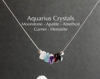 May include: A delicate necklace featuring a string of colorful gemstones, including moonstone, apatite, amethyst, garnet, and hematite. The necklace is labeled "Aquarius Crystals" with the names of the stones. The gemstones are in shades of white, blue, purple, red, and black.