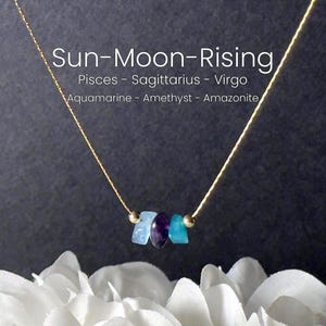 May include: A gold necklace with three gemstones in shades of blue and purple. The necklace is set against a dark backdrop, with the text "Sun-Moon-Rising" and astrological signs. The necklace is displayed over white flowers.