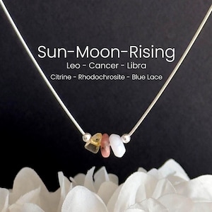 May include: A silver necklace with three gemstones in yellow, pink, and white. The necklace has the text "Sun-Moon-Rising" and astrological signs. The gemstones are citrine, rhodochrosite, and blue lace. The necklace is displayed against a dark background.