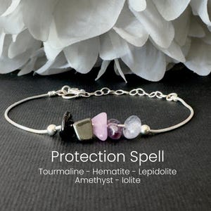 May include: A silver bracelet with a series of colorful stones. The stones include black tourmaline, hematite, pink lepidolite, amethyst, and iolite. The text "Protection Spell" is below the bracelet.