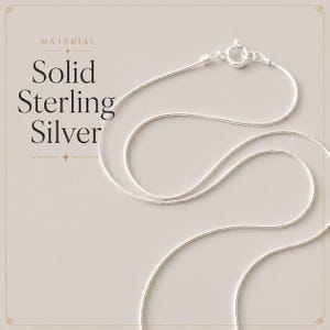May include: A thin, flexible silver snake chain necklace featuring a circular spring ring clasp. The text on the left side reads: MATERIAL Solid Sterling Silver. The chain is arranged in a curved, looping pattern against a neutral background.