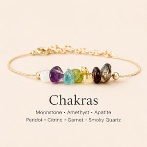 Può includere: Un braccialetto con catena dorata con sette pietre preziose colorate: pietra di luna, ametista, apatite, peridoto, citrino, granato e quarzo fumé. La parola "Chakras" è stampata sotto.