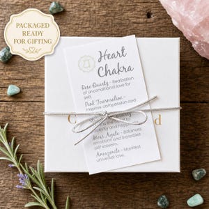 May include: A white gift box tied with a silver ribbon, with a tag that reads "Heart Chakra." The tag lists the properties of Rose Quartz, Pink Tourmaline, Star Quartz, Moss Agate, and Amazonite. The box is surrounded by small green stones.