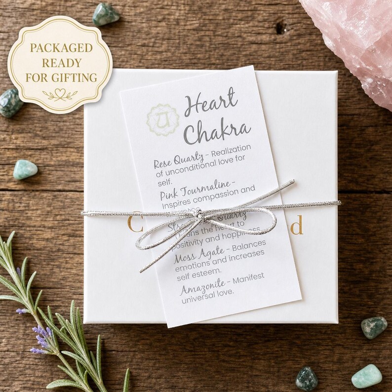 May include: A white gift box tied with a silver ribbon, with a tag that reads "Heart Chakra." The tag lists the properties of Rose Quartz, Pink Tourmaline, Star Quartz, Moss Agate, and Amazonite. The box is surrounded by small green stones.