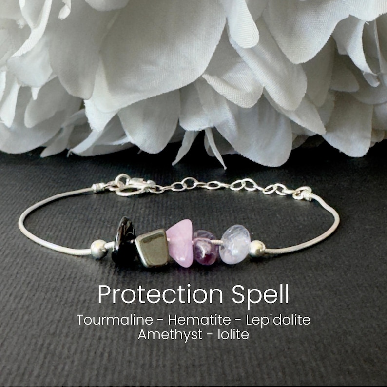 May include: A silver bracelet with a series of colorful stones. The stones include black tourmaline, hematite, pink lepidolite, amethyst, and iolite. The text "Protection Spell" is below the bracelet.